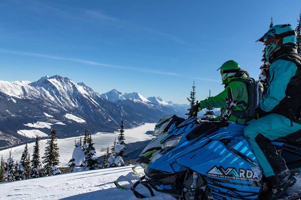 Two snowmobilers looking out over mountain and lake view